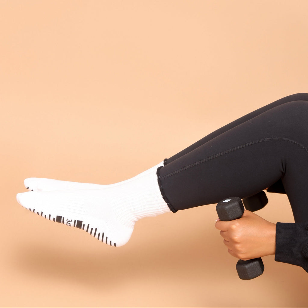 A person wearing black leggings and white socks lifts a small black dumbbell while seated against a solid warm beige background.