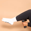 A person wearing black leggings and white socks lifts a small black dumbbell while seated against a solid warm beige background.
