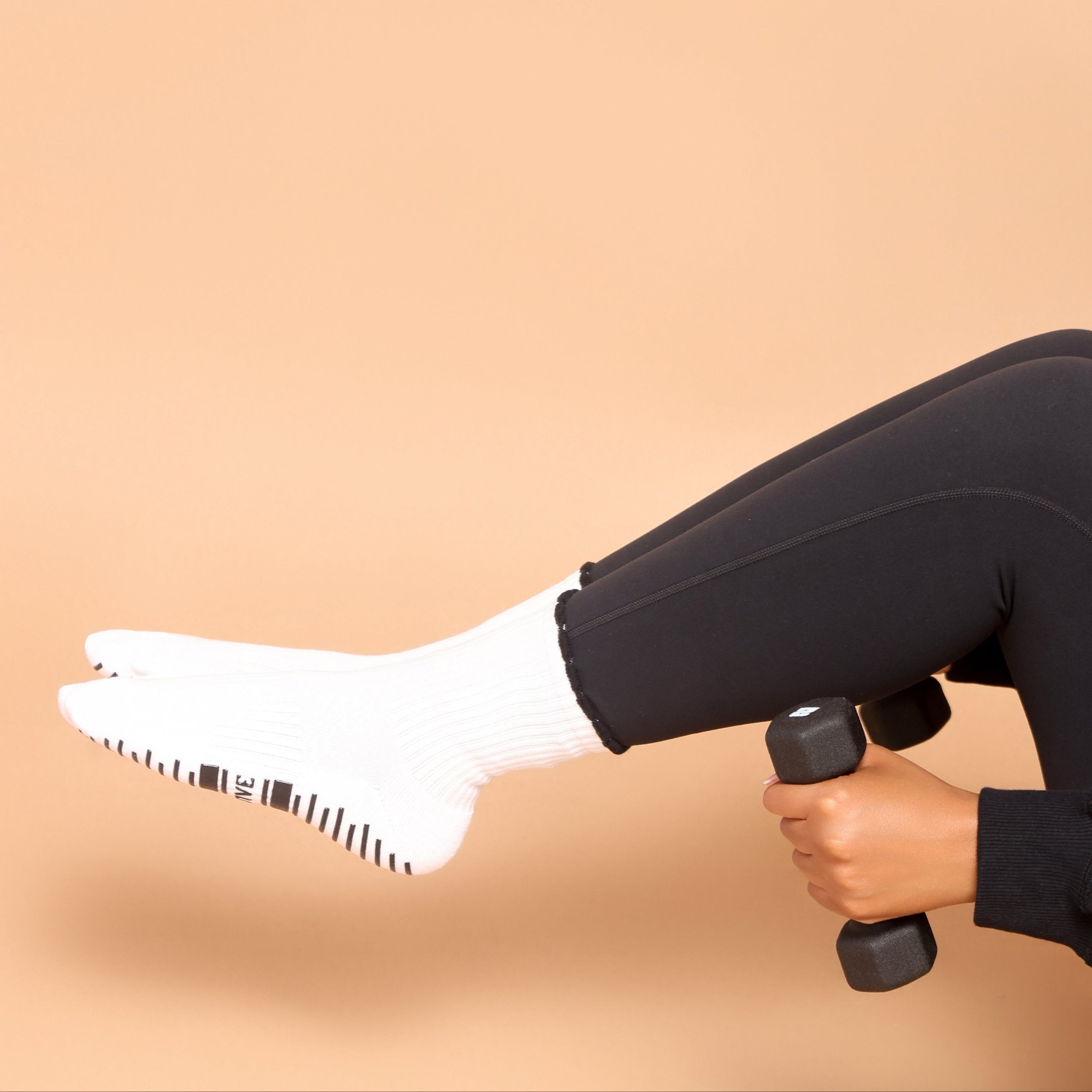 A person wearing black leggings and white socks lifts a small black dumbbell while seated against a solid warm beige background.